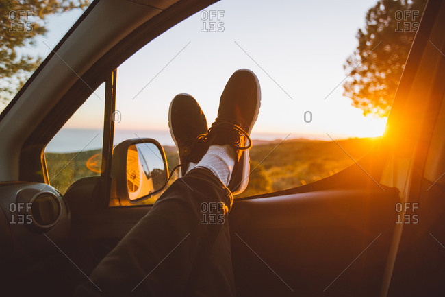 Legs of anonymous man lying on car window against view of magnificent countryside and bright setting sun
