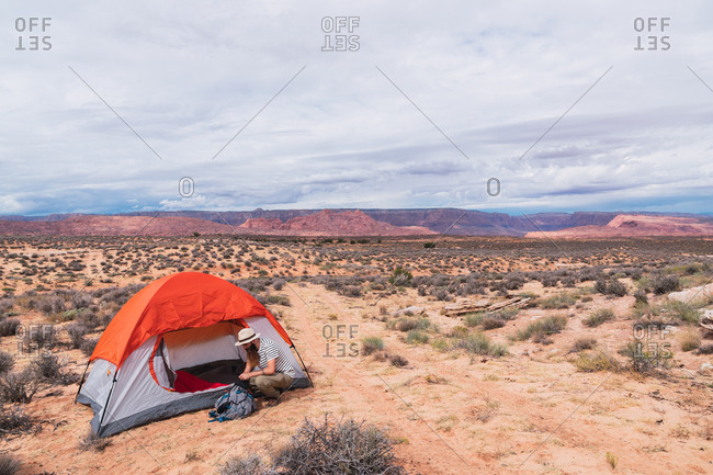 Side view of handsome guy in casual outfit packing backpack while sitting on ground near tent on camping area in desert