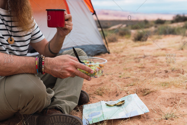 Crop man eating salad and enjoying hot drink while sitting on sandy ground near map and compass during camping in desert