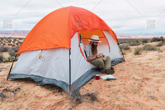 man eating salad and enjoying hot drink while sitting on sandy ground near map and compass during camping in desert