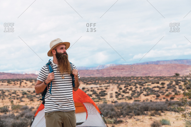 Handsome bearded man with backpack looking away while walking near tent on cloudy day in desert