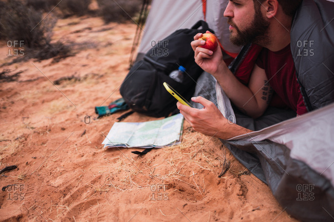 Crop bearded man eating fresh apple and browsing modern smartphone while lying in tent during camping in desert