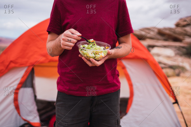 Crop man with bowl of fresh salad standing near modern tent on camping area in desert