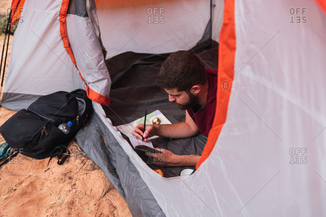 From above shot of young guy lying in tent and drawing in sketchbook while camping in desert