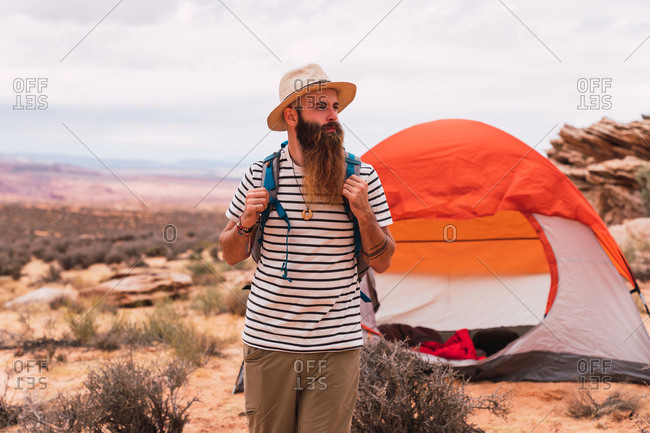 Handsome bearded guy with backpack looking away while standing on blurred background of amazing desert