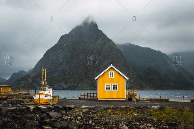 Landscape of little yellow house standing on coast of blue river on background of high mountains covered with fog