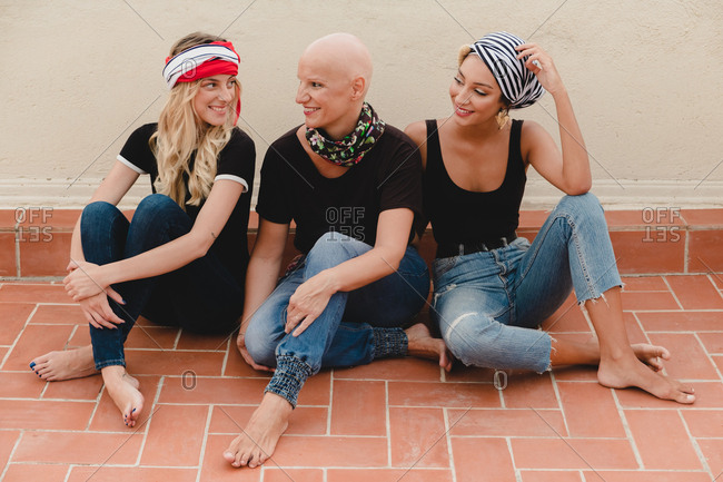 Three barefoot women in black T-shirts and jeans sitting on tile floor near wall and smiling