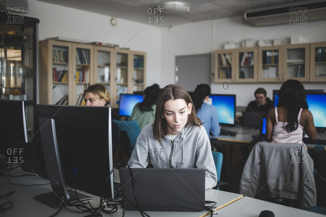 Confident female high school student using laptop at desk against teacher and friends sitting in computer lab