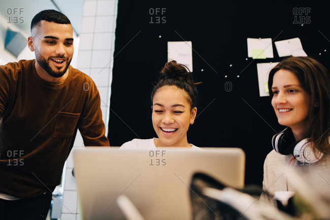 Smiling young businesswoman discussing with multi-ethnic colleagues over laptop against bulletin board at office