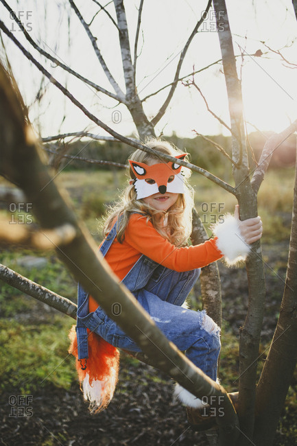 Portrait of girl in fox costume sitting on bare tree against sky during Halloween at sunset