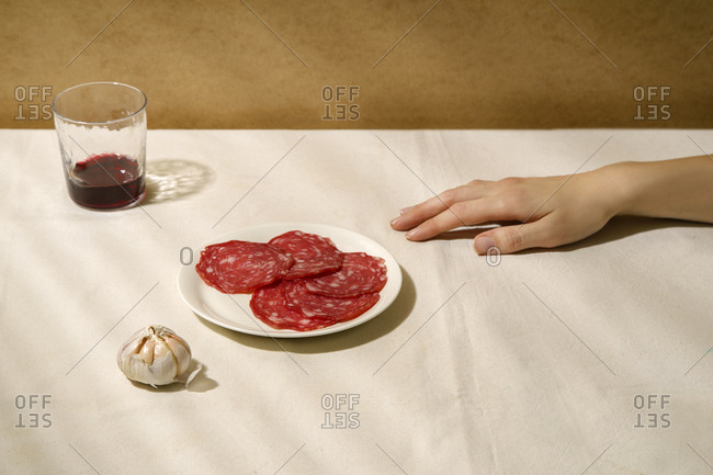 Minimalistic composition with salami slices on a white plate, garlic, a glass of wine and female hand