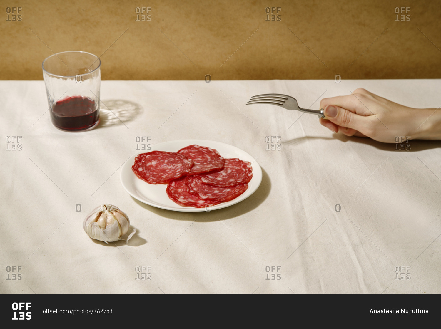 Minimalistic composition with salami slices on a white plate, garlic, a glass of wine and female hand holding fork