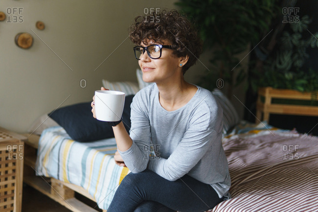 Woman with curly hair wearing retro glasses drinking coffee on bed