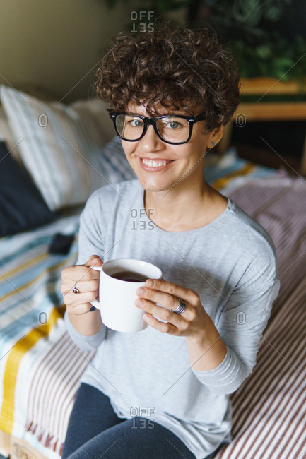 Smiling woman with curly hair wearing retro glasses drinking coffee on bed