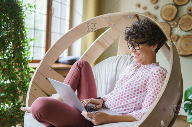Woman with curly hair laughing while sitting in wooden round chair using laptop
