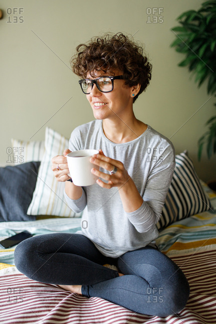 Happy woman wearing retro glasses drinking coffee on bed
