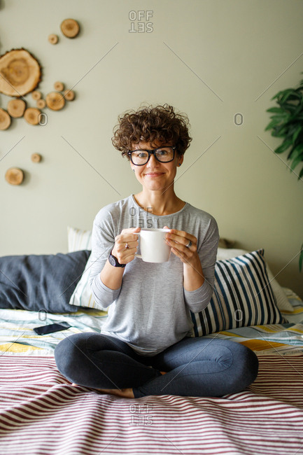 Smiling woman wearing retro glasses drinking coffee on bed