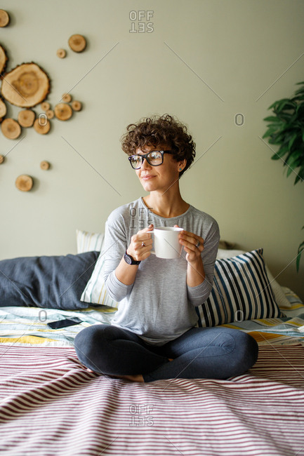 Woman wearing retro glasses drinking coffee on bed