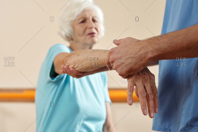 Unrecognizable physician exercising hand of defocused elderly Caucasian woman wincing at pain while getting physical therapy in recovery center