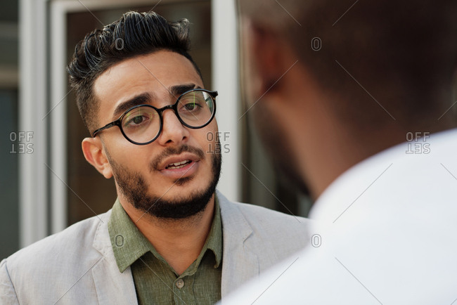 Waist-up portrait of young good-looking Middle Eastern bearded man in glasses  talking to unrecognizable male colleague outdoors