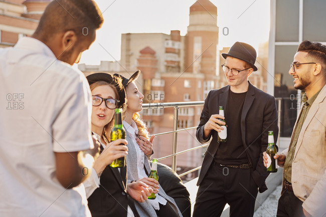 Multiethnic group of young fashionable male and female students standing on rooftop cafe terrace, chatting and drinking beer