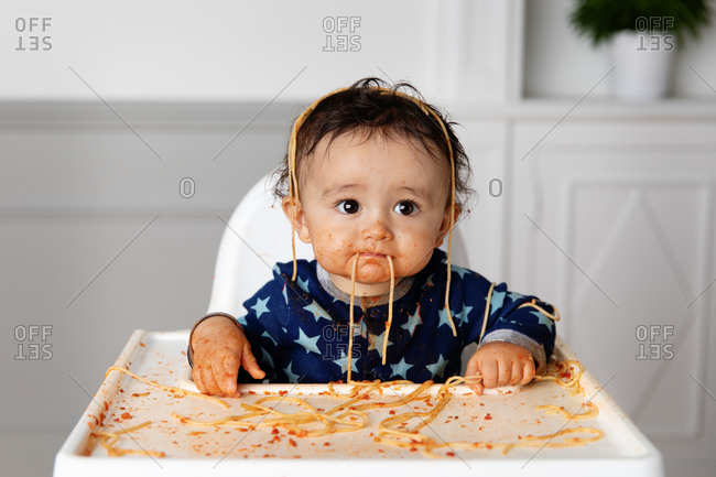 Frowning toddler in high chair making a mess while eating spaghetti