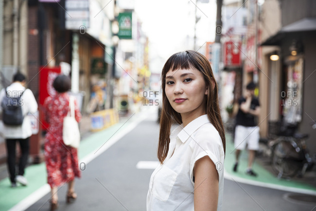Smiling Japanese woman with long brown hair wearing white short-sleeved blouse standing in a street.