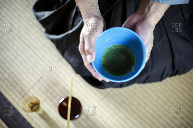 High angle close up of Japanese man wearing traditional kimono kneeling on floor holding blue bowl with Matcha tea during tea ceremony.