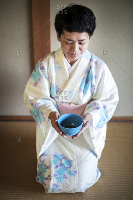 Japanese woman wearing traditional white kimono with blue floral pattern kneeling on floor during tea ceremony, holding blue tea bowl.