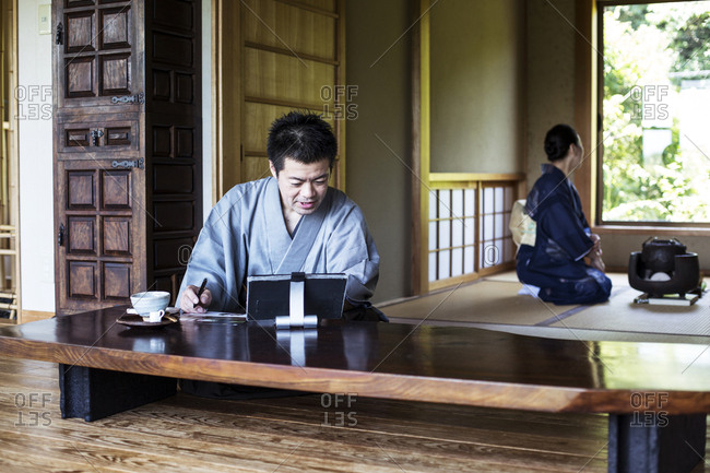 Japanese man wearing kimono sitting on floor in traditional Japanese house, looking at digital tablet.