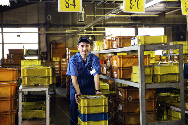 Japanese man wearing baseball cap and blue overall standing in factory, yellow crates on shelves.