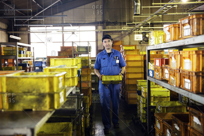 Japanese man wearing baseball cap and blue overall standing in factory, holding yellow plastic crate.