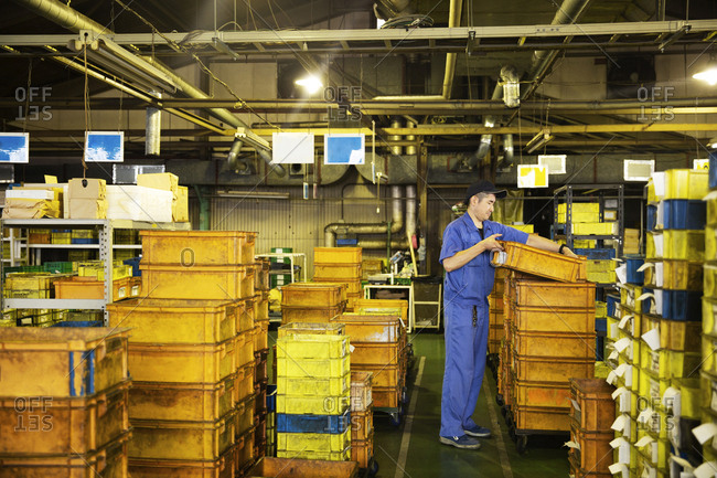 Japanese man wearing baseball cap and blue overall standing in factory, stacking orange plastic crates.