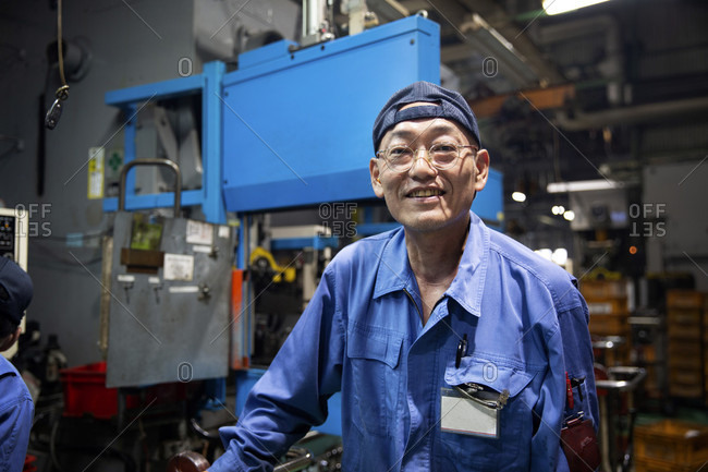 Japanese man wearing glasses and blue overall standing in factory, smiling at camera.