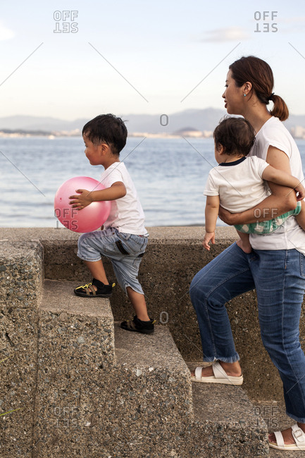 Japanese woman carrying toddler and young boy with pink ball walking up concrete steps by the ocean.