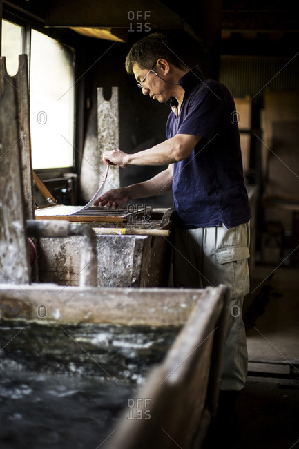 Japanese man in a workshop, holding a wooden frame with pressed pulp, making traditional Washi paper.