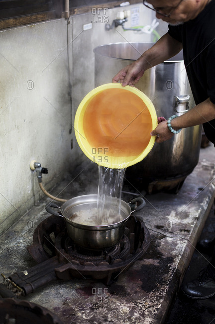 Japanese man standing in a textile plant dye workshop, pouring water from yellow plastic bucket into pot.