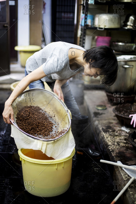 Japanese woman standing in a textile plant dye workshop, pouring plant dye from a sieve into a yellow bucket..
