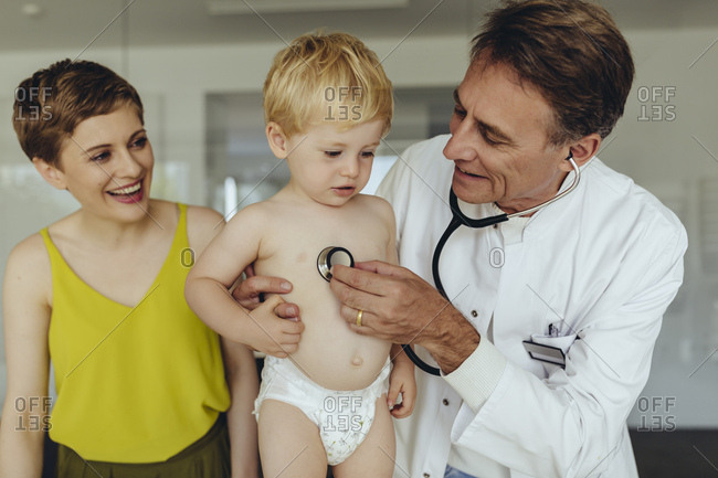 Pediatrician examining toddler with stethoscope- mother standing next to them