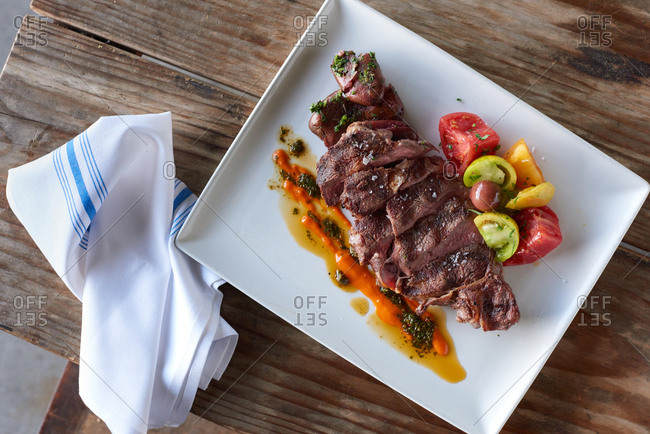 A grilled steak, sliced on a plate presented with a small side of seasoned heirloom tomatoes, a roasted red pepper aioli and a classic chimichurri sauce, on a square plate on a rustic wood table in a modern bistro restaurant.