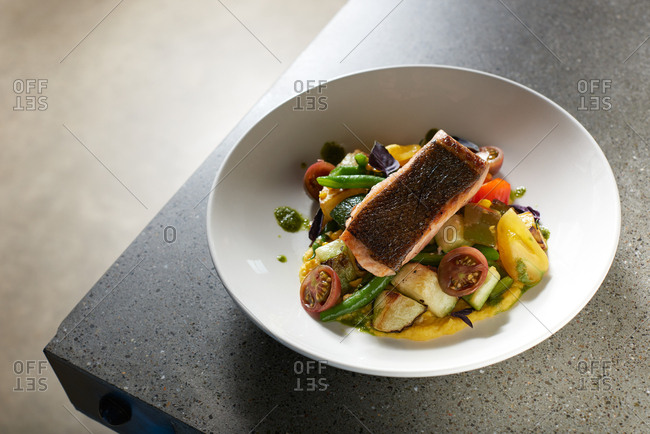 Pan Fried King Salmon served skin side up on a bed of summer vegetables including heirloom cherry tomatoes, green beans, and eggplant served on creamy polenta in a white bowl on a modern concrete surface.