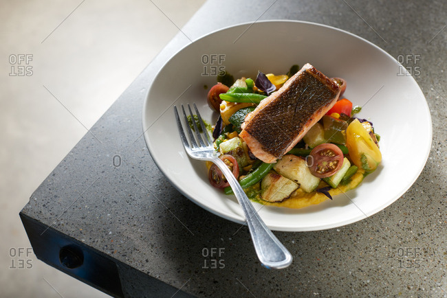 Pan Fried King Salmon served skin side up on a bed of summer vegetables including heirloom cherry tomatoes, green beans, and eggplant served on creamy polenta in a white bowl on a modern concrete surface.