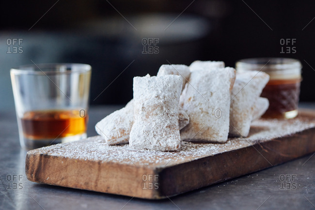 A thick wooden board holds a half dozen small beignets, with a small jar of bourbon caramel sauce in the back, with a glass of bourbon straight up sits nearby.