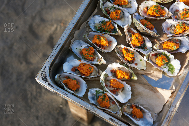 Oysters prepped in a large hotel pan with compound butter, chopped bacon and chives, ready to be grilled outdoors.