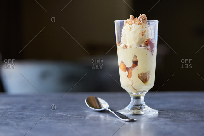 A single serving portion of peach trifle consisting of cooked peaches, custard, chunks of cake and vanilla ice cream, presented in a tall glass served in a farm to table restaurant.