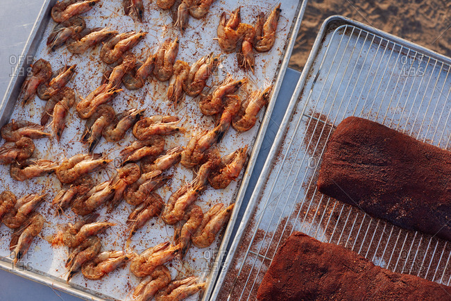 Two trays being prepped for outdoor cooking, one containing seasoned raw shrimp still in the shell and the other with two racks of rib with rub on them.