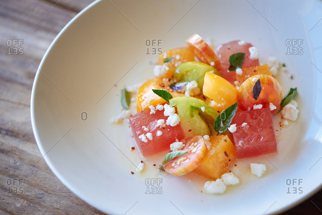 A heirloom tomato salad with feta cheese, watermelon and mint, drizzled with olive oil and presented in a shallow white bowl at a farm to table restaurant.