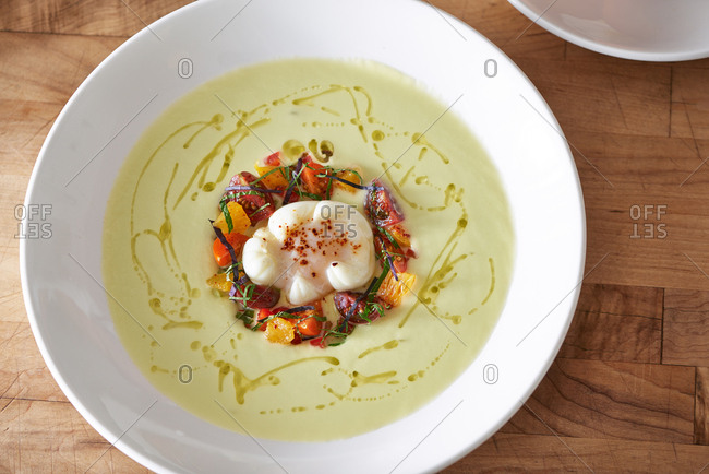 A small arrangement of cucumbers, citrus, heirloom tomato, basil strips, poblano peppers holding an olive oil poached egg with dried red pepper on top.  It is surrounded by a cucumber puree with drizzled olive oil in the bowl.