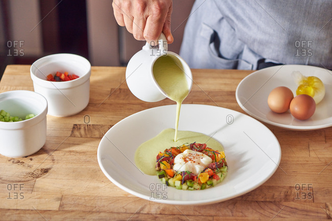 A small arrangement of cucumbers, citrus, heirloom tomato, basil strips, poblano peppers holding an olive oil poached egg with dried red pepper on top.  A chef pours cucumber puree into the bowl with various mis en place remaining in place.