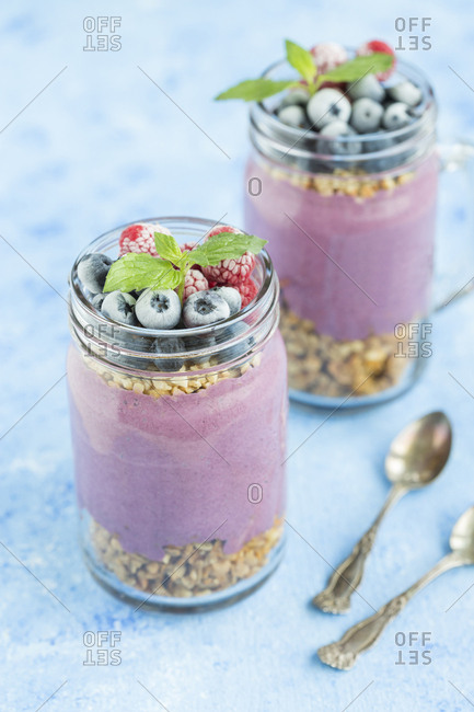 Two glasses of yoghurt with peanut granola- aronia powder and topping of chopped hazelnuts and frozen berries